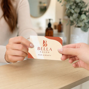 Premium Glossy Business Cards in use - being handed across a light wooden salon reception counter from one hand to another